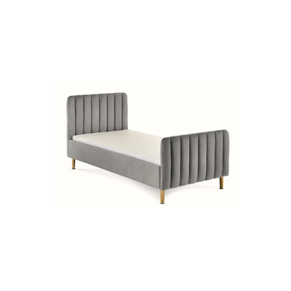A gray velvet single bed with a tufted headboard and rolled footboard, styled in a bedroom with patterned wallpaper, a white dresser, a floor lamp, and a light blue throw blanket.