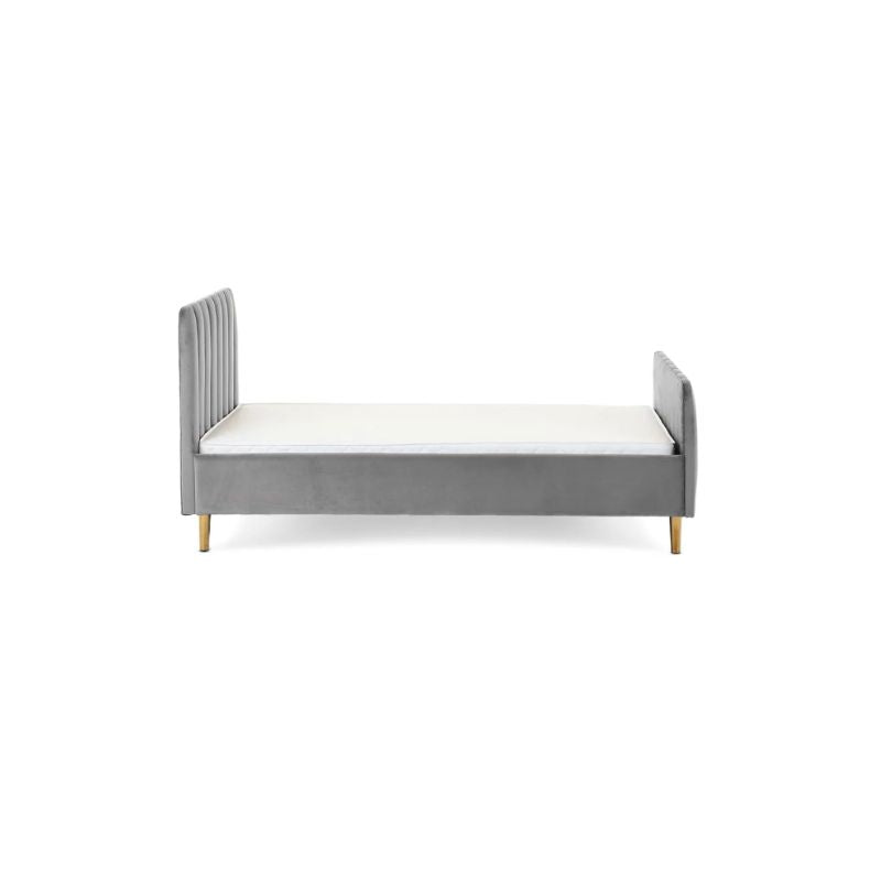 A gray velvet single bed with a tufted headboard and rolled footboard, styled in a bedroom with patterned wallpaper, a white dresser, a floor lamp, and a light blue throw blanket.