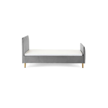 A gray velvet single bed with a tufted headboard and rolled footboard, styled in a bedroom with patterned wallpaper, a white dresser, a floor lamp, and a light blue throw blanket.