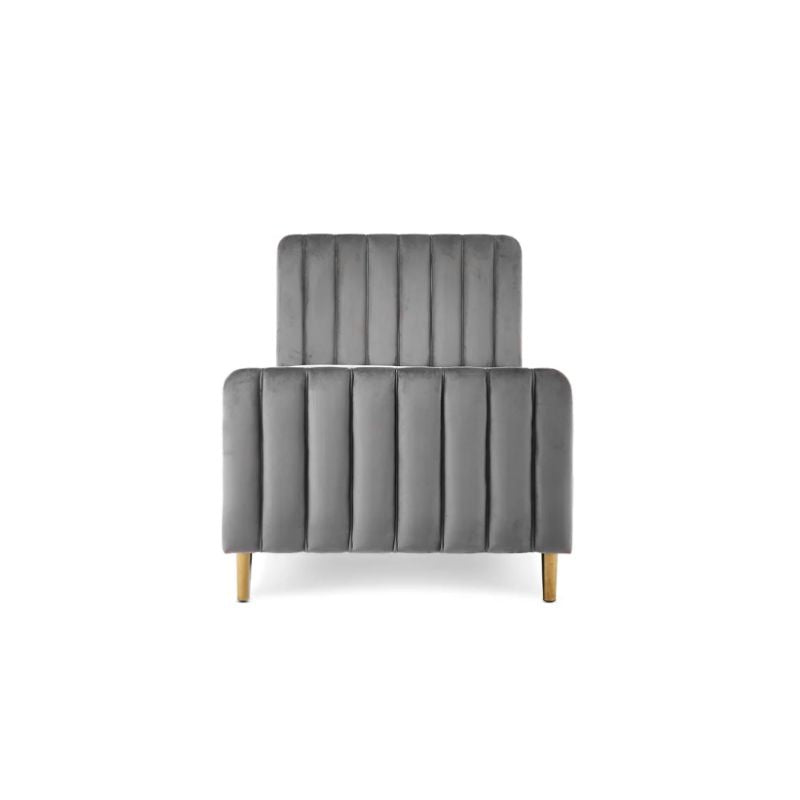 A gray velvet single bed with a tufted headboard and rolled footboard, styled in a bedroom with patterned wallpaper, a white dresser, a floor lamp, and a light blue throw blanket.