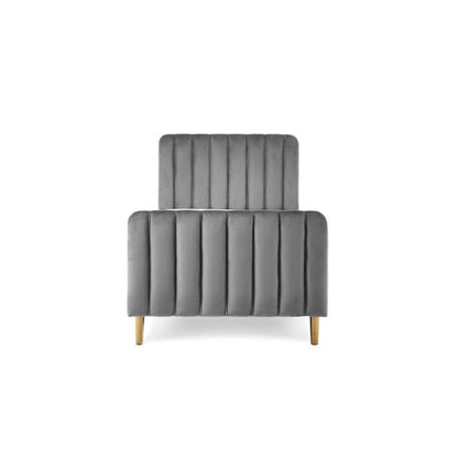 A gray velvet single bed with a tufted headboard and rolled footboard, styled in a bedroom with patterned wallpaper, a white dresser, a floor lamp, and a light blue throw blanket.