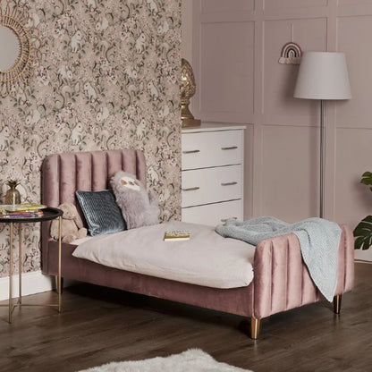 A pink velvet single bed with gold legs in a styled bedroom setting, featuring a patterned wallpaper background, a white dresser, a floor lamp, and a side table.