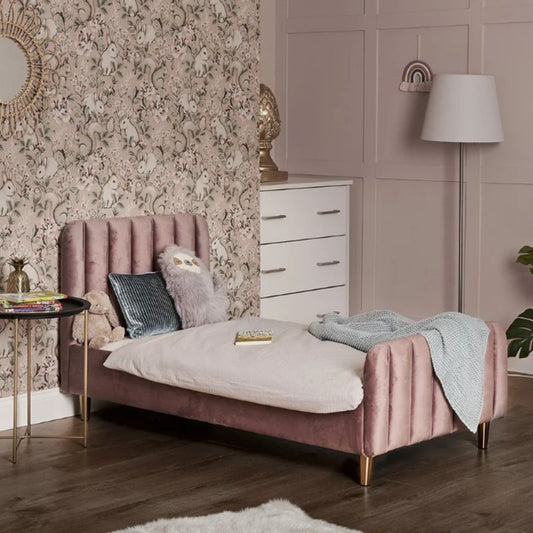 A pink velvet single bed with gold legs in a styled bedroom setting, featuring a patterned wallpaper background, a white dresser, a floor lamp, and a side table.