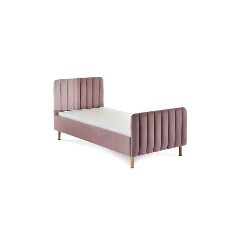 A pink velvet single bed with gold legs in a styled bedroom setting, featuring a patterned wallpaper background, a white dresser, a floor lamp, and a side table.