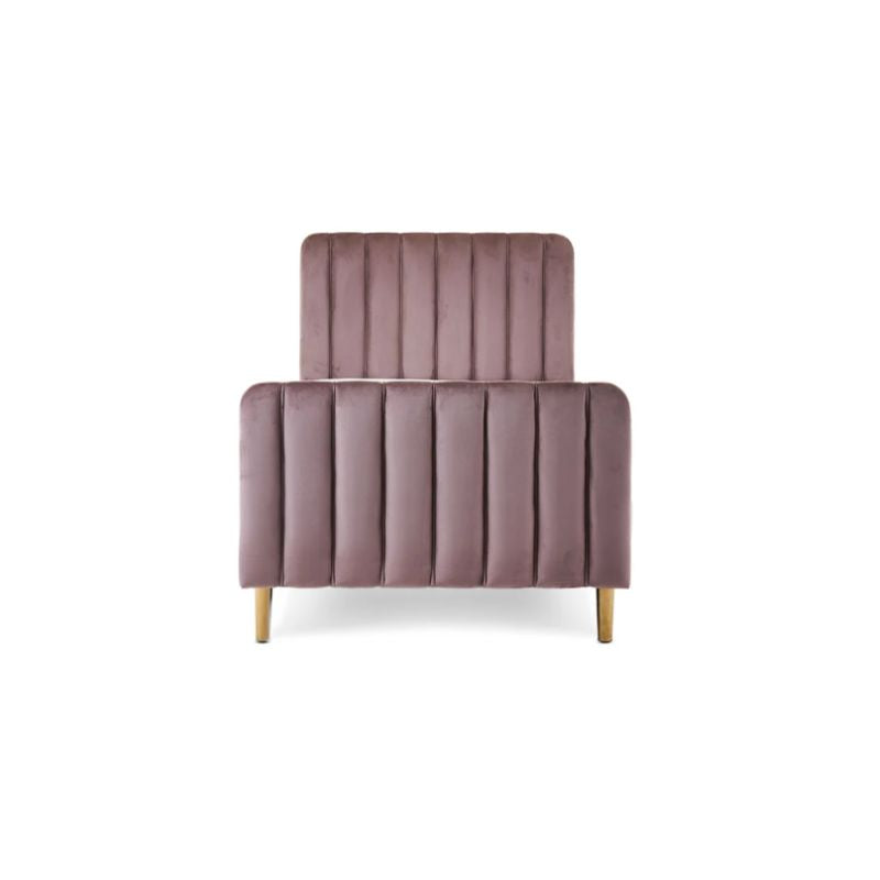 A pink velvet single bed with gold legs in a styled bedroom setting, featuring a patterned wallpaper background, a white dresser, a floor lamp, and a side table.