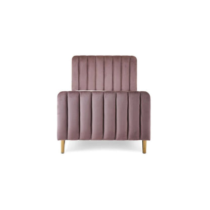 A pink velvet single bed with gold legs in a styled bedroom setting, featuring a patterned wallpaper background, a white dresser, a floor lamp, and a side table.