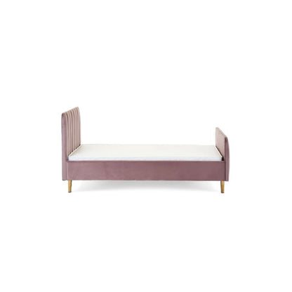 A pink velvet single bed with gold legs in a styled bedroom setting, featuring a patterned wallpaper background, a white dresser, a floor lamp, and a side table.