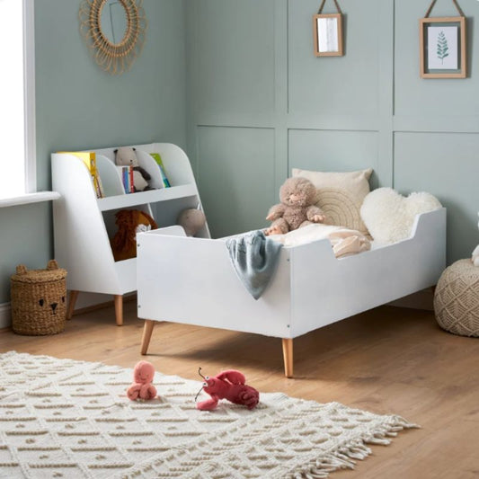 A white wooden toddler bed with light wood legs and a curved headboard, styled in a nursery with a white bookshelf, soft toys, a textured rug, and pale blue paneled walls.