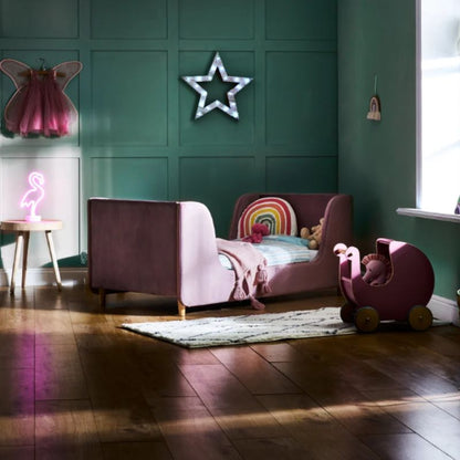 A pink velvet children's daybed in a styled nursery room with dark green paneled walls, a neon flamingo lamp, a star light, and a pink toy stroller on the wooden floor.