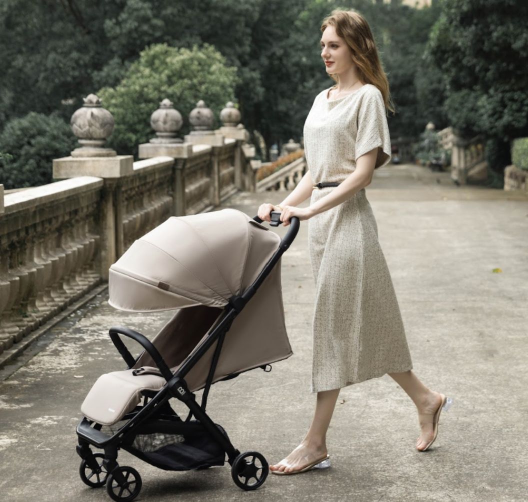 A compact stroller with a light beige (latte) canopy and seat liner, a black frame, and black wheels, shown from a three-quarters angle on a solid white background.