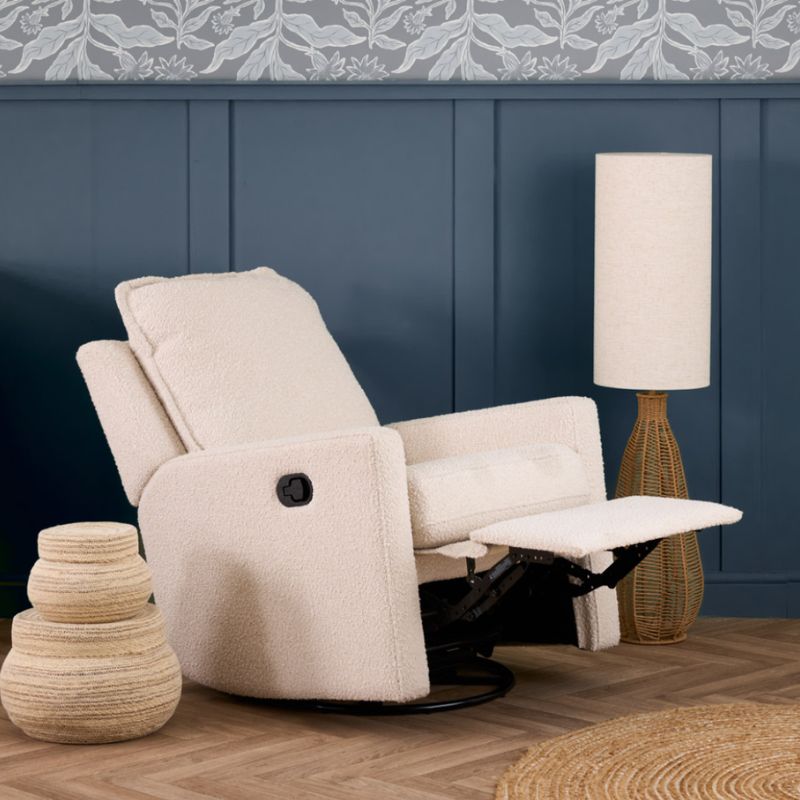 An upholstered recliner chair in a textured cream bouclé fabric, shown with the footrest extended in a styled room setting with a blue paneled wall, a floor lamp, and a decorative basket.