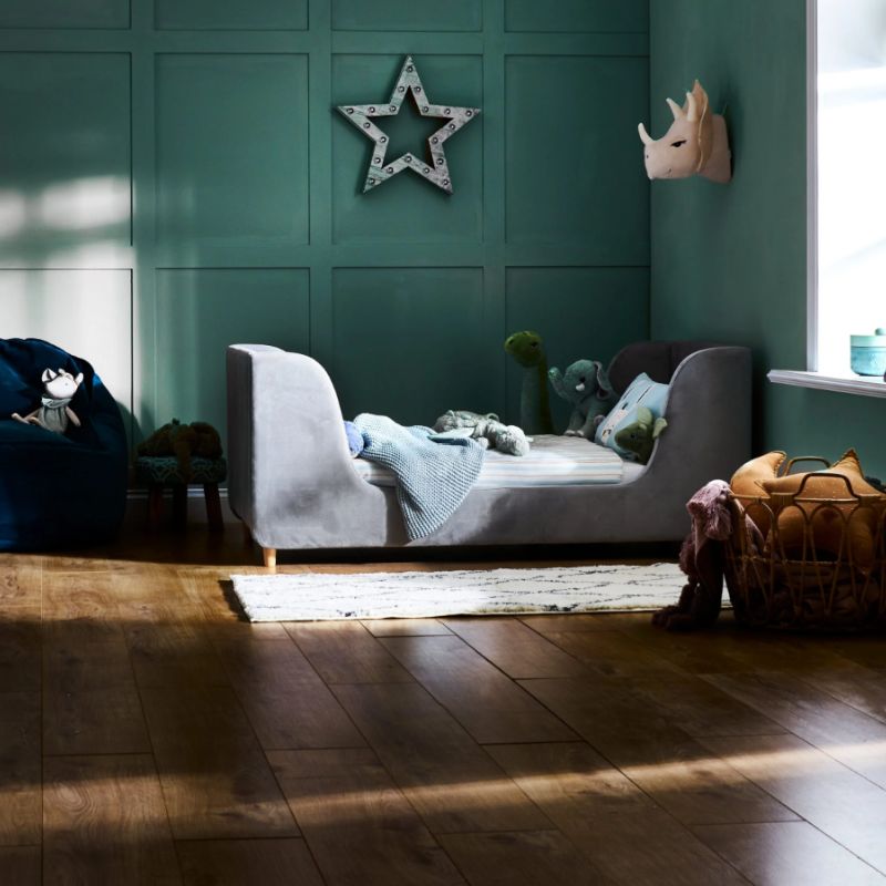 A gray upholstered children's daybed in a styled nursery or playroom with teal paneled walls, a star wall decor piece, a window with bright sunlight, a bean bag, and a basket of toys.